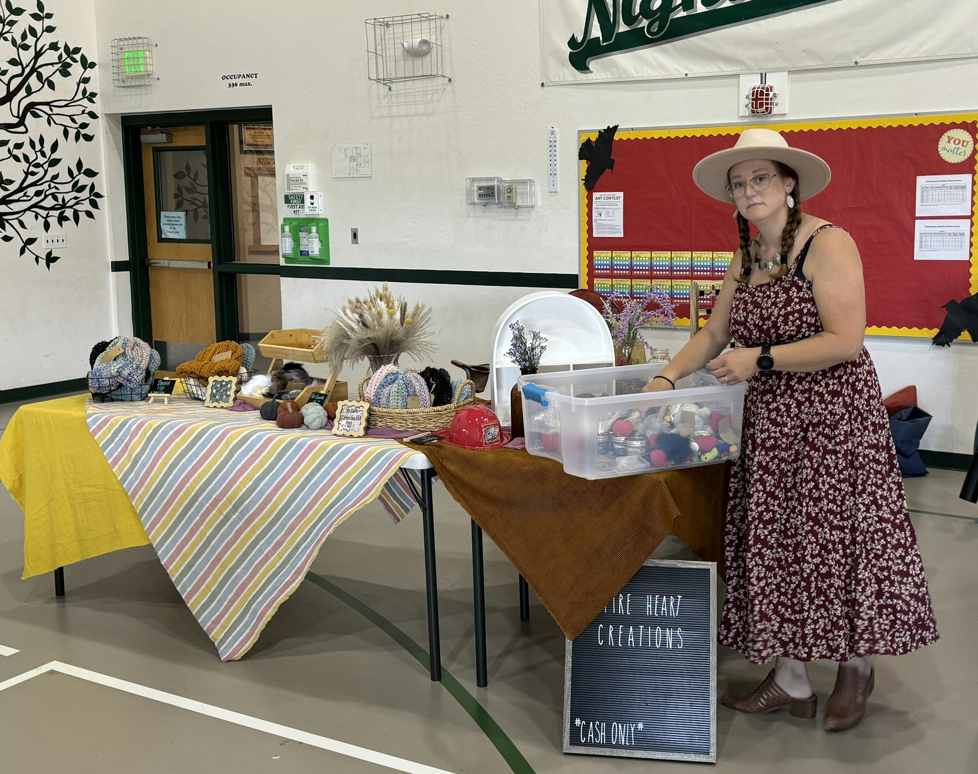 A woman at a craft stall with hats, dried flowers, and a sign saying "Cash Only."