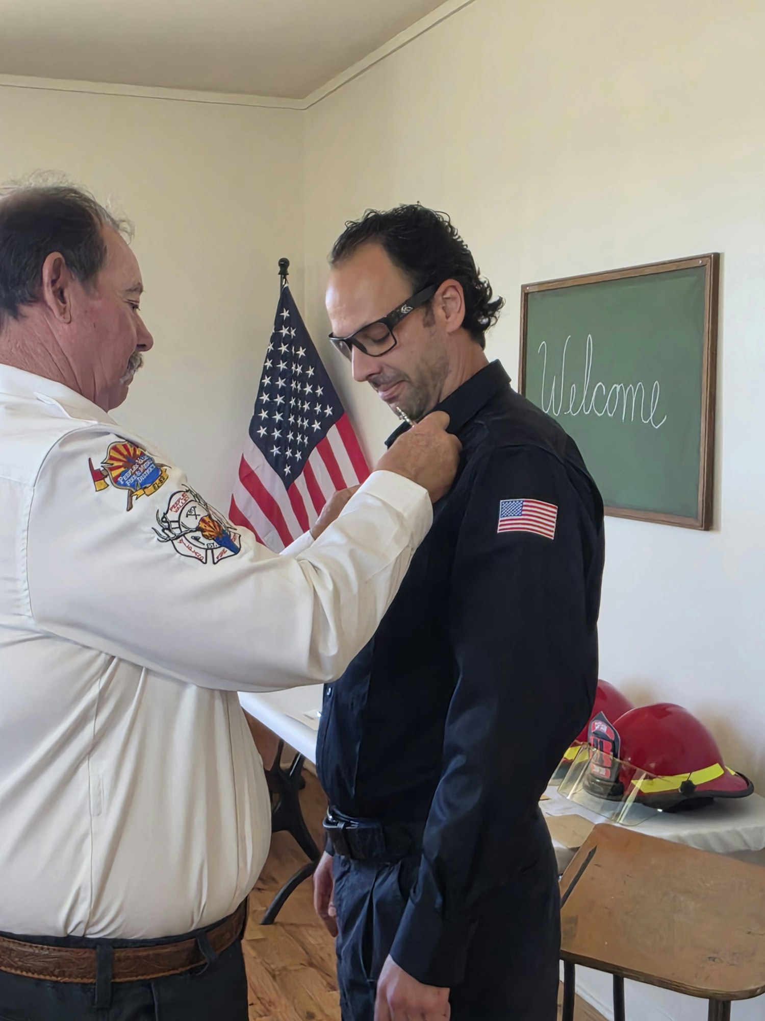 A man is being pinned or honored by another person in front of an American flag, possibly at a ceremony or welcome event.