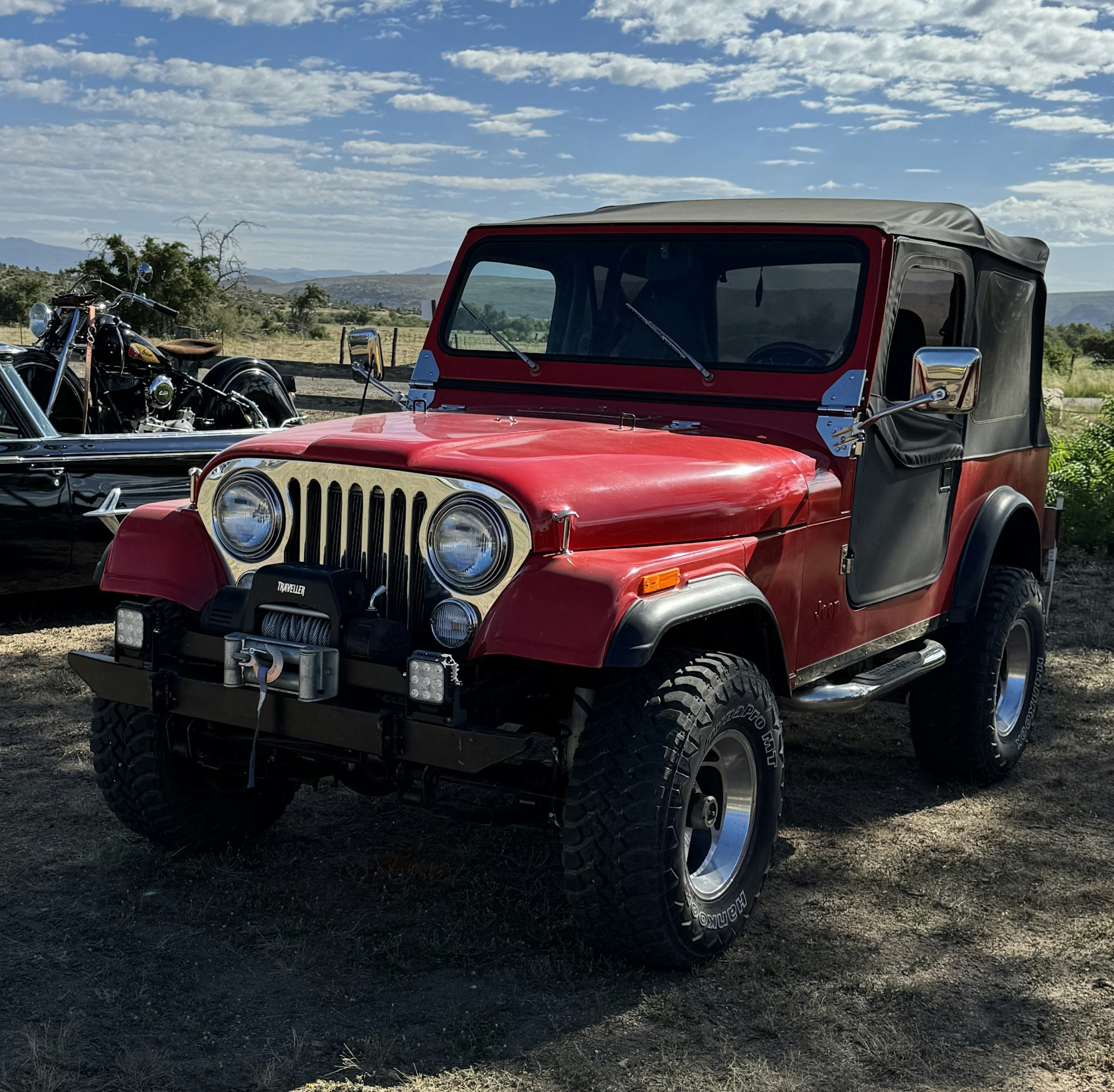 A red vintage Jeep with a soft top parked in a field with a motorcycle and trailer in the background.