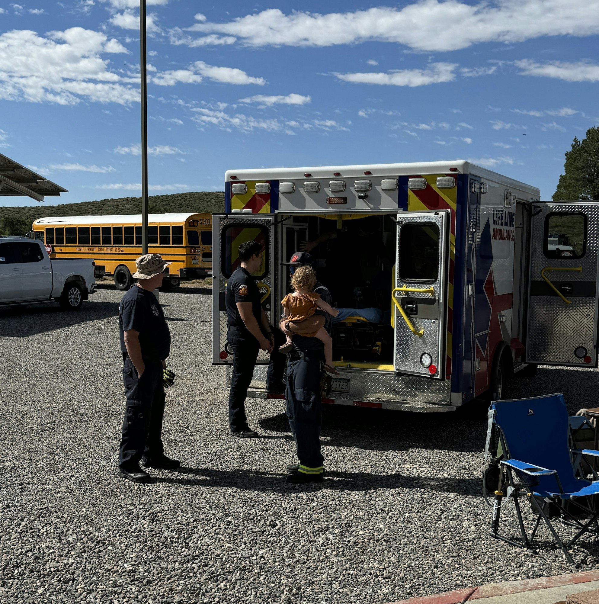 Ambulance with open doors, two emergency responders, a child, and parked vehicles under a clear sky.