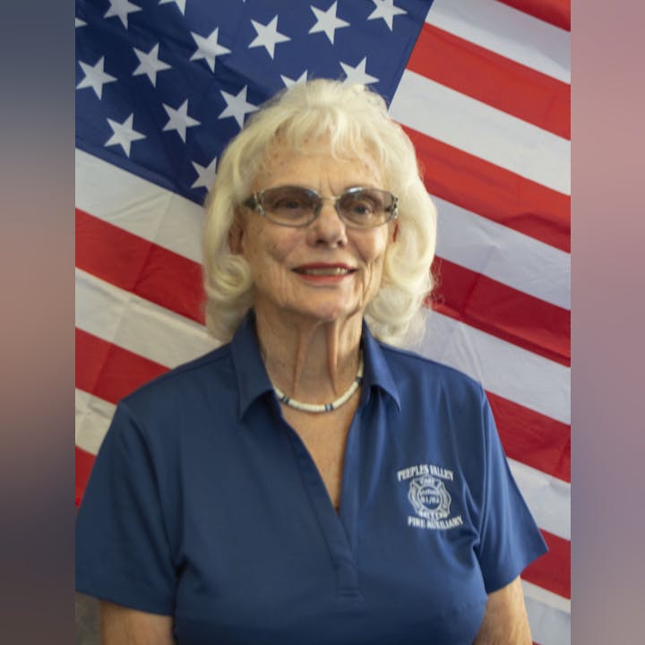 An older woman in a blue shirt stands in front of an American flag background, smiling at the camera.