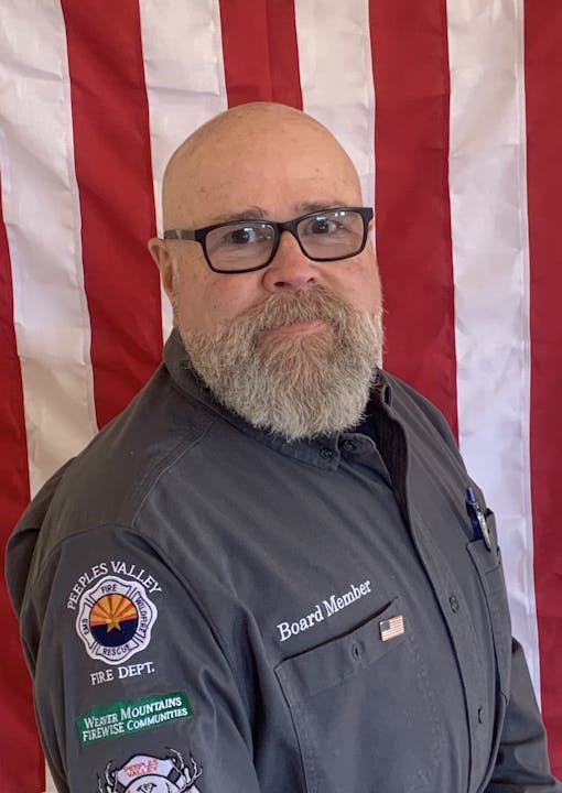 A man in glasses with a beard stands in front of an American flag, wearing a fire department uniform with multiple patches and "Board Member" text.