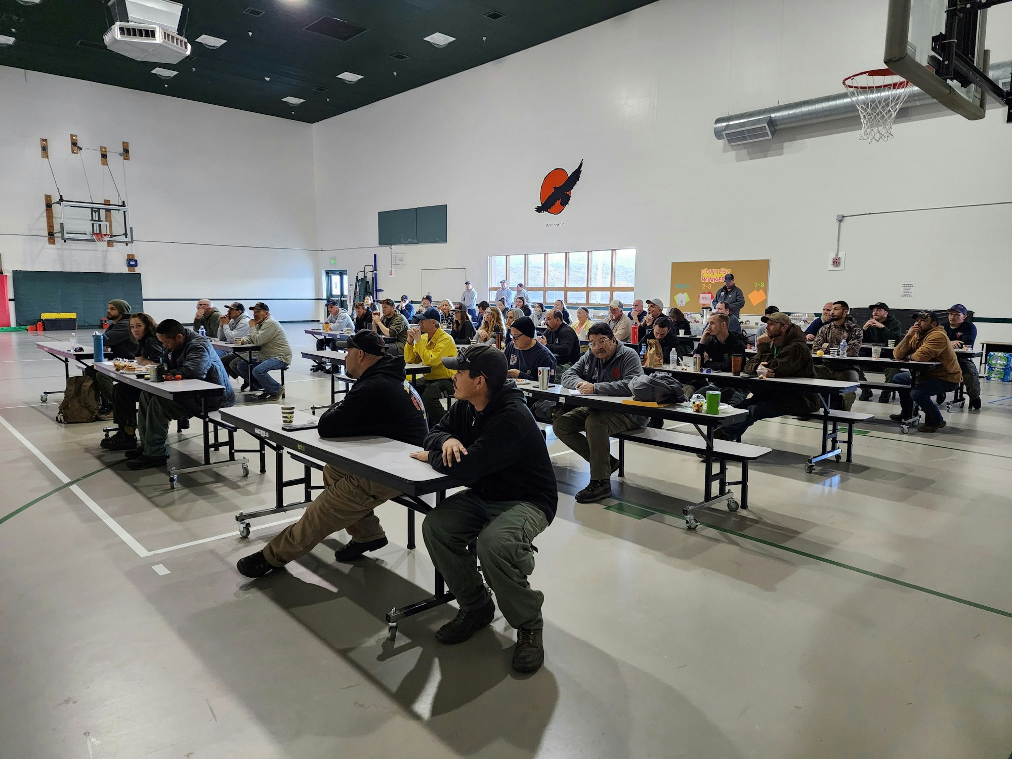 A group of people sitting at long tables in a gymnasium or multi-purpose room, possibly engaged in a meeting or event.