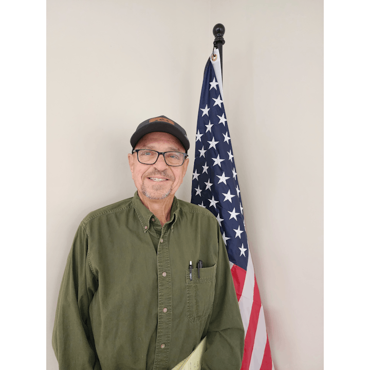 A man wearing glasses and a cap stands in front of a U.S. flag.