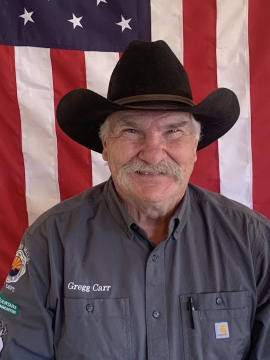 A man in a cowboy hat stands in front of an American flag, wearing a shirt with "Gregg Carr" embroidered on it.