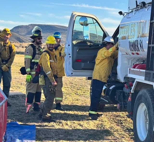 A group of firefighters in protective gear are gathered around a fire truck, preparing for a response in a rural area.