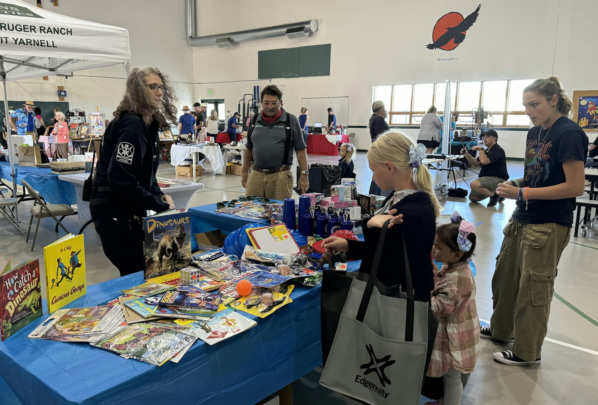 Indoor market with people browsing items on tables, such as books and toys, with a "Welcome" sign in the background.