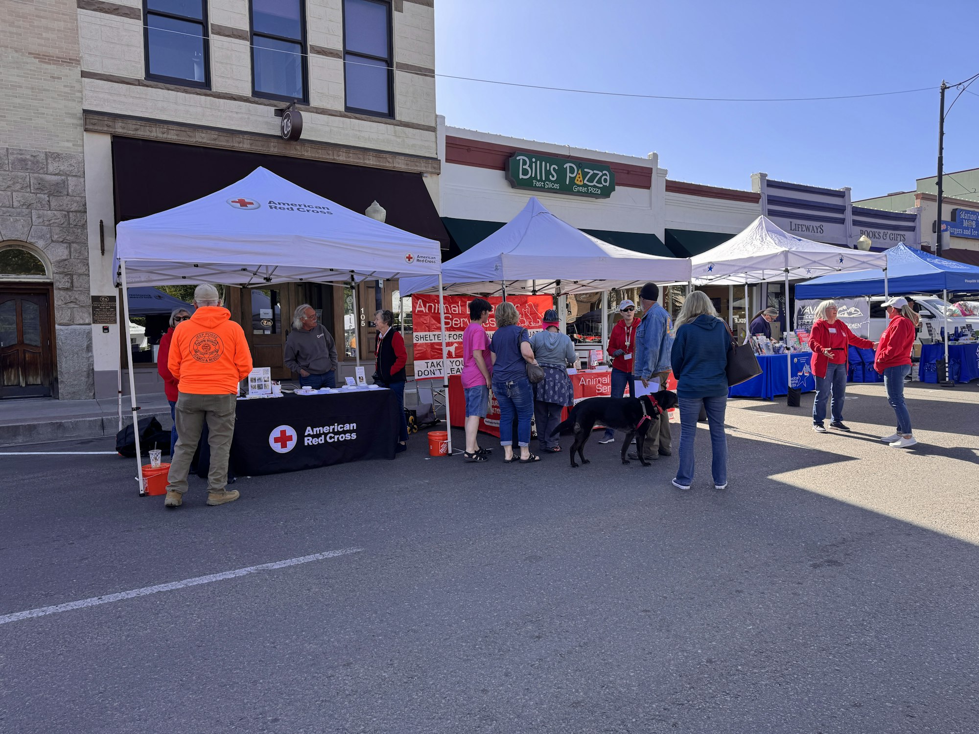 A street event featuring tents, including an American Red Cross booth, with people engaging and a dog present.