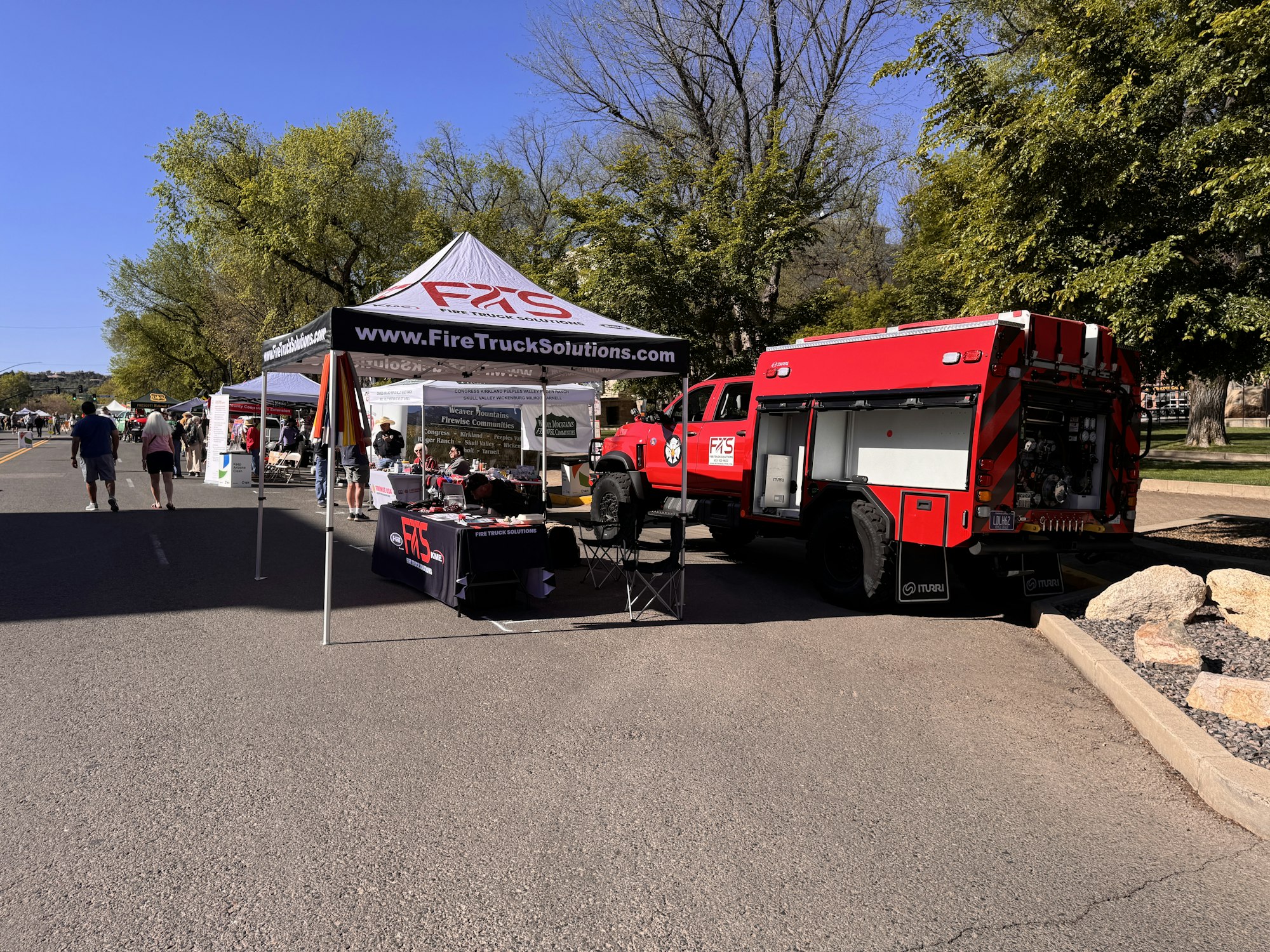 A street scene featuring vendor tents, a red fire truck, and people browsing at an outdoor event. Clear blue sky above.