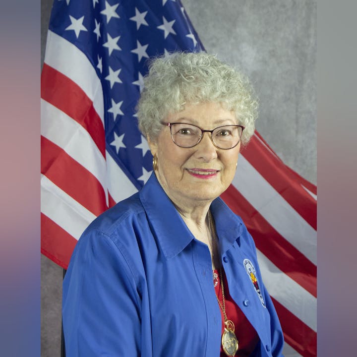 An elderly woman with glasses, smiling, in a blue shirt, wearing a medal, in front of the American flag.