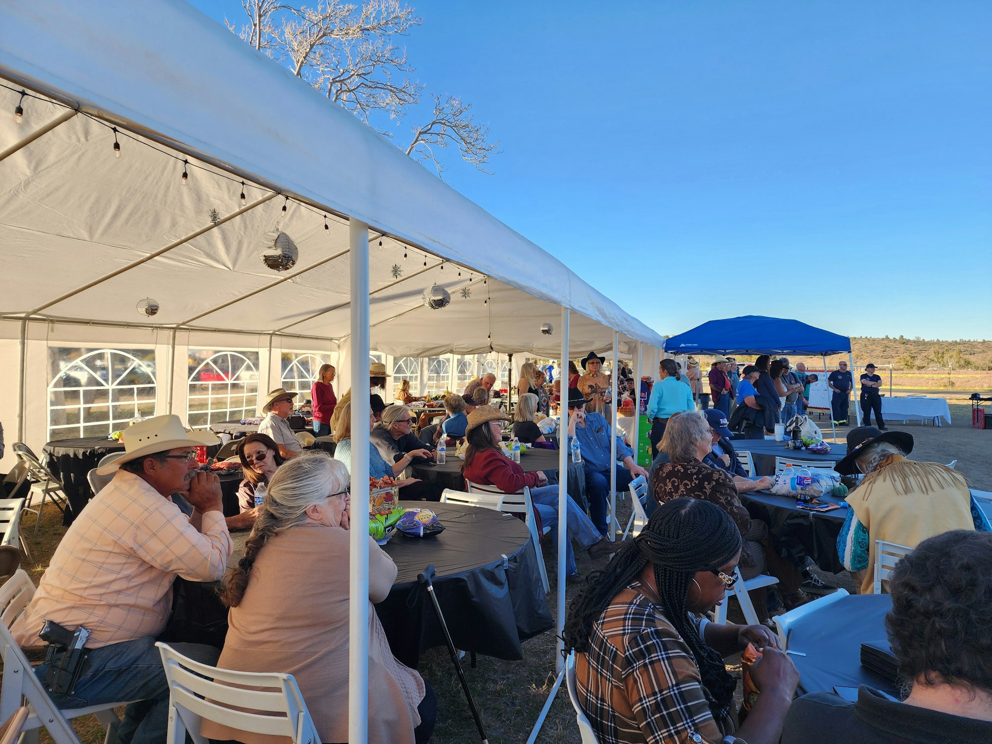 People are seated under a large white tent at an outdoor gathering, socializing and enjoying the event.