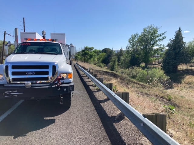 A tow truck parked on a roadside with guardrails, clear skies, and trees in the background.