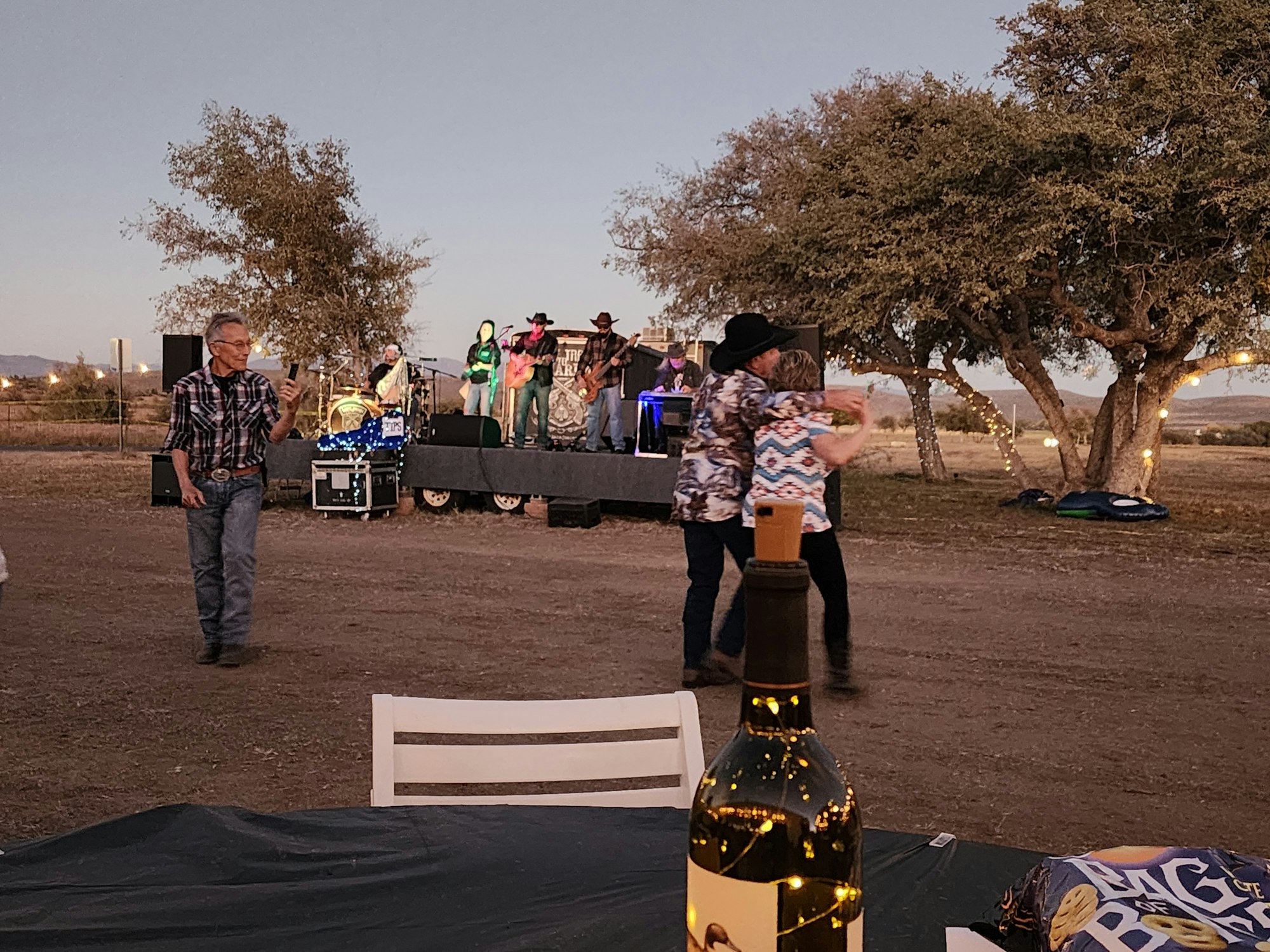 Outdoor party with a live band, people dancing, and a bottle on a table in the foreground.