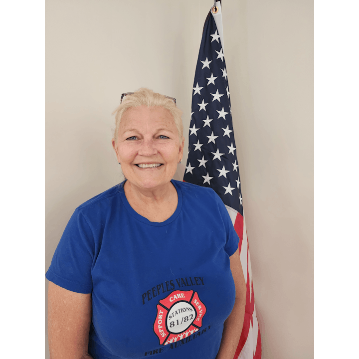 A person smiling, wearing a blue shirt with a fire department logo, in front of an American flag.