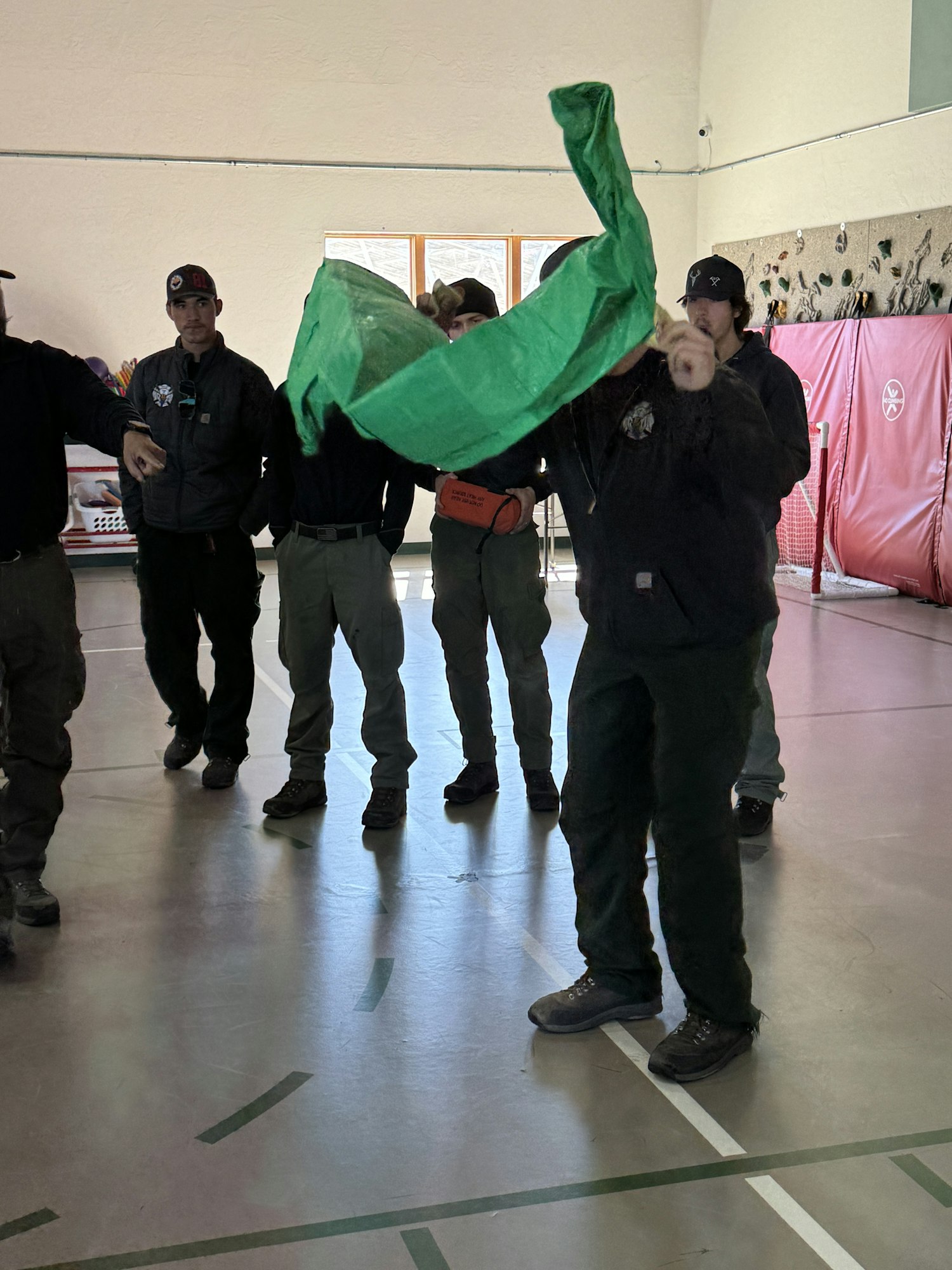 Group of people in uniform, one holding a green tarp or cloth, indoors on a gym floor.