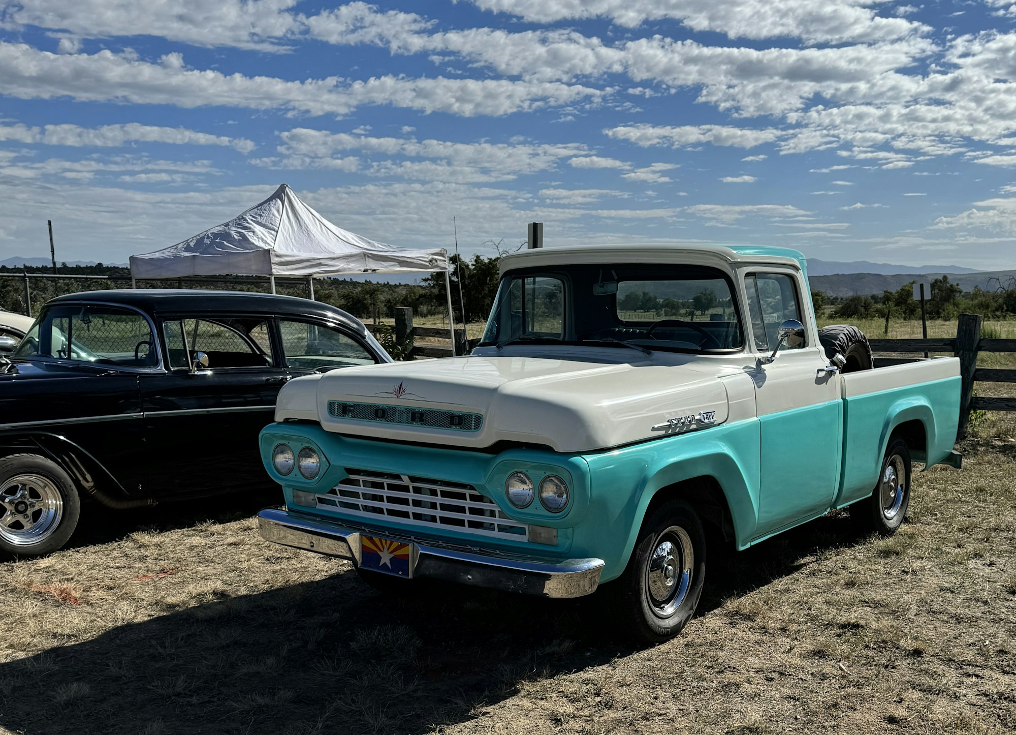 A two-tone vintage pickup truck and a classic car at an outdoor event with a tent and mountains in the background.