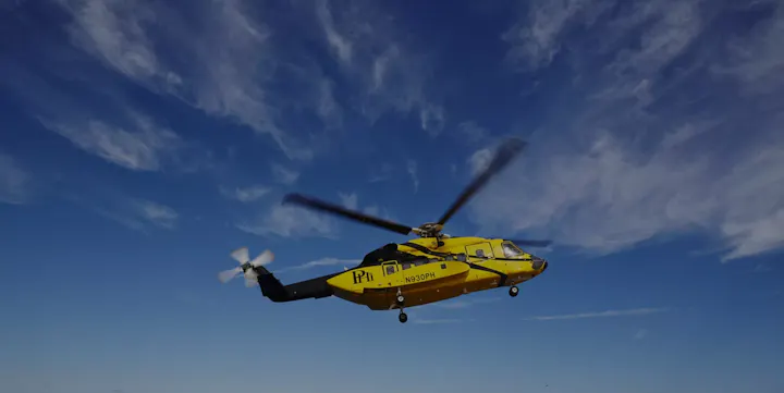 A yellow helicopter is flying against a blue sky with scattered clouds. The rotor blades are in motion.