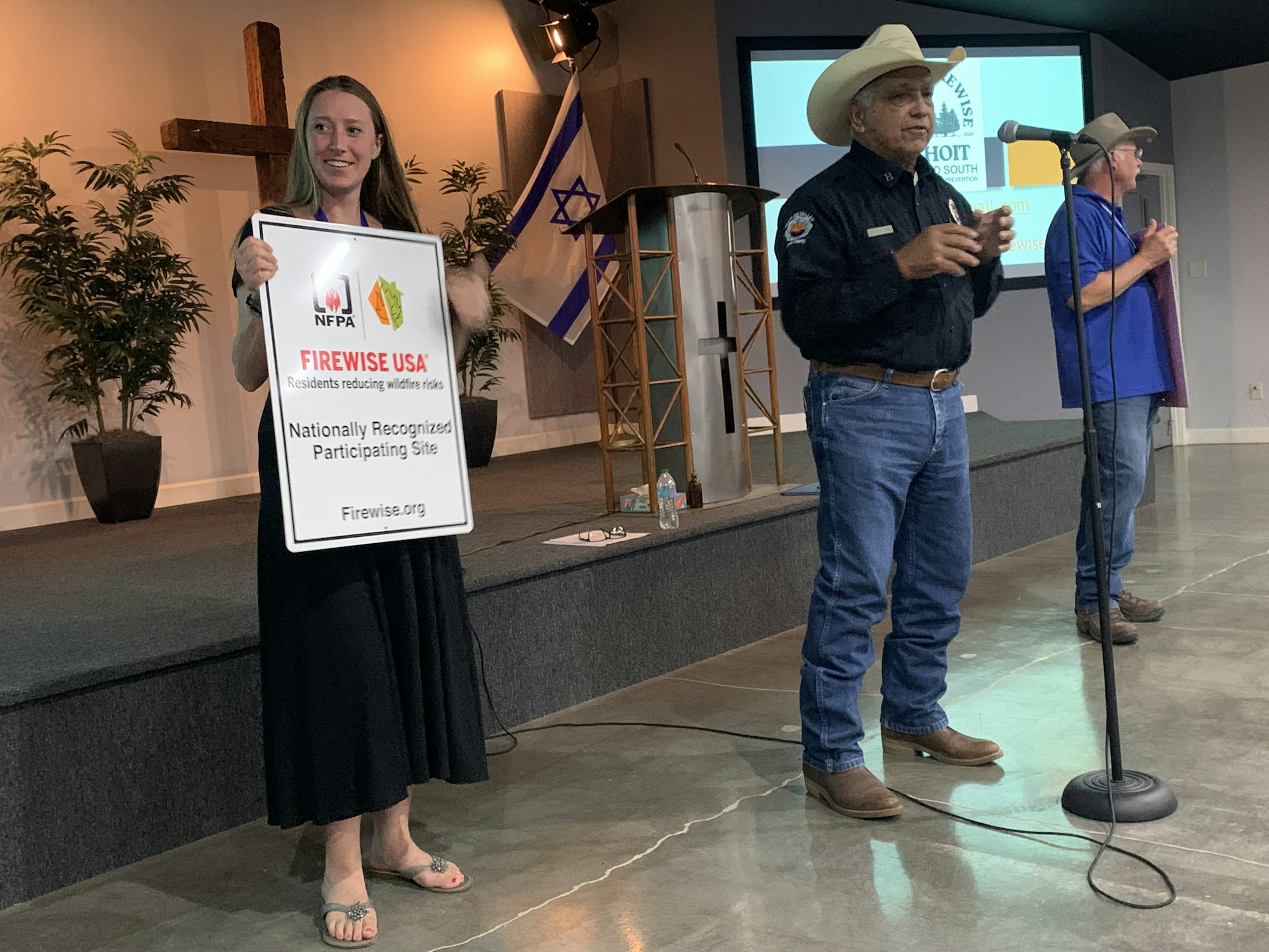 Three people are on a stage, one holding a Firewise USA sign, with a presentation setup and flags in the background.