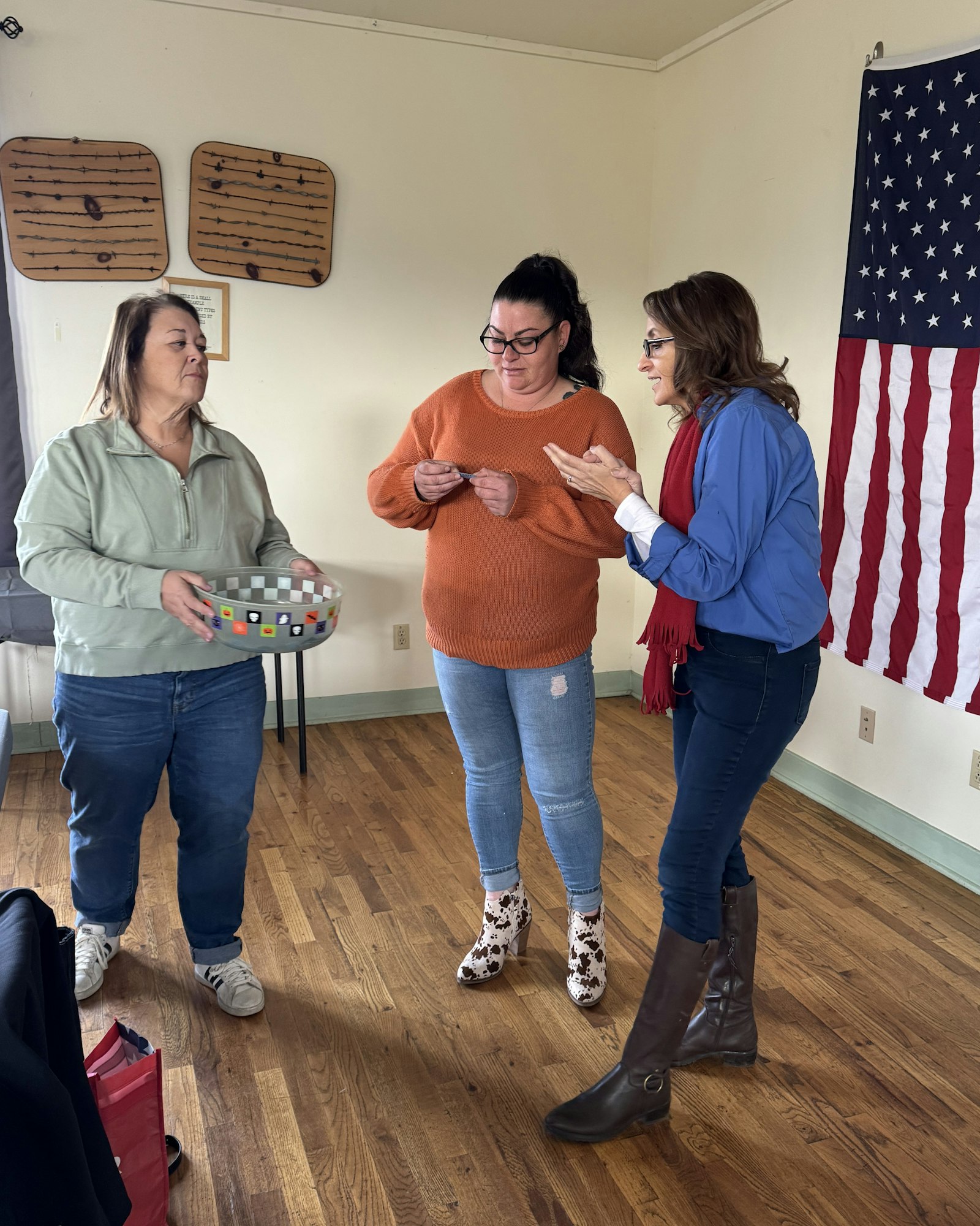 Three women interacting, one holding a bowl and papers, in a room with an American flag and key holders on the wall.