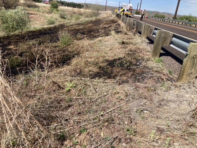 Dry grassy roadside with wooden fence posts and a vehicle with yellow lights in the distance.