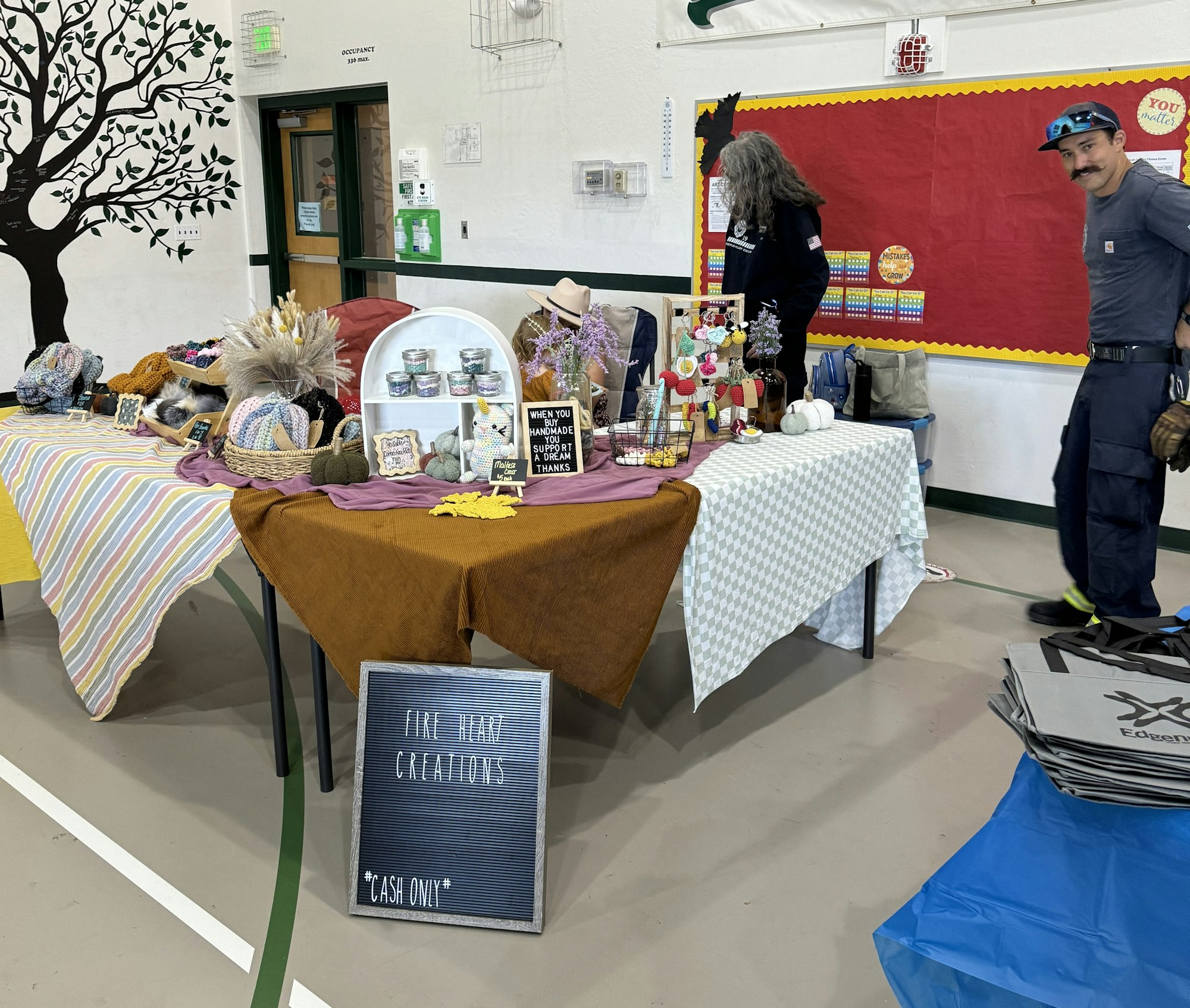 A craft fair with handmade items on tables and two people. Sign reads "FINE HAIR CREATIONS - CASH ONLY."