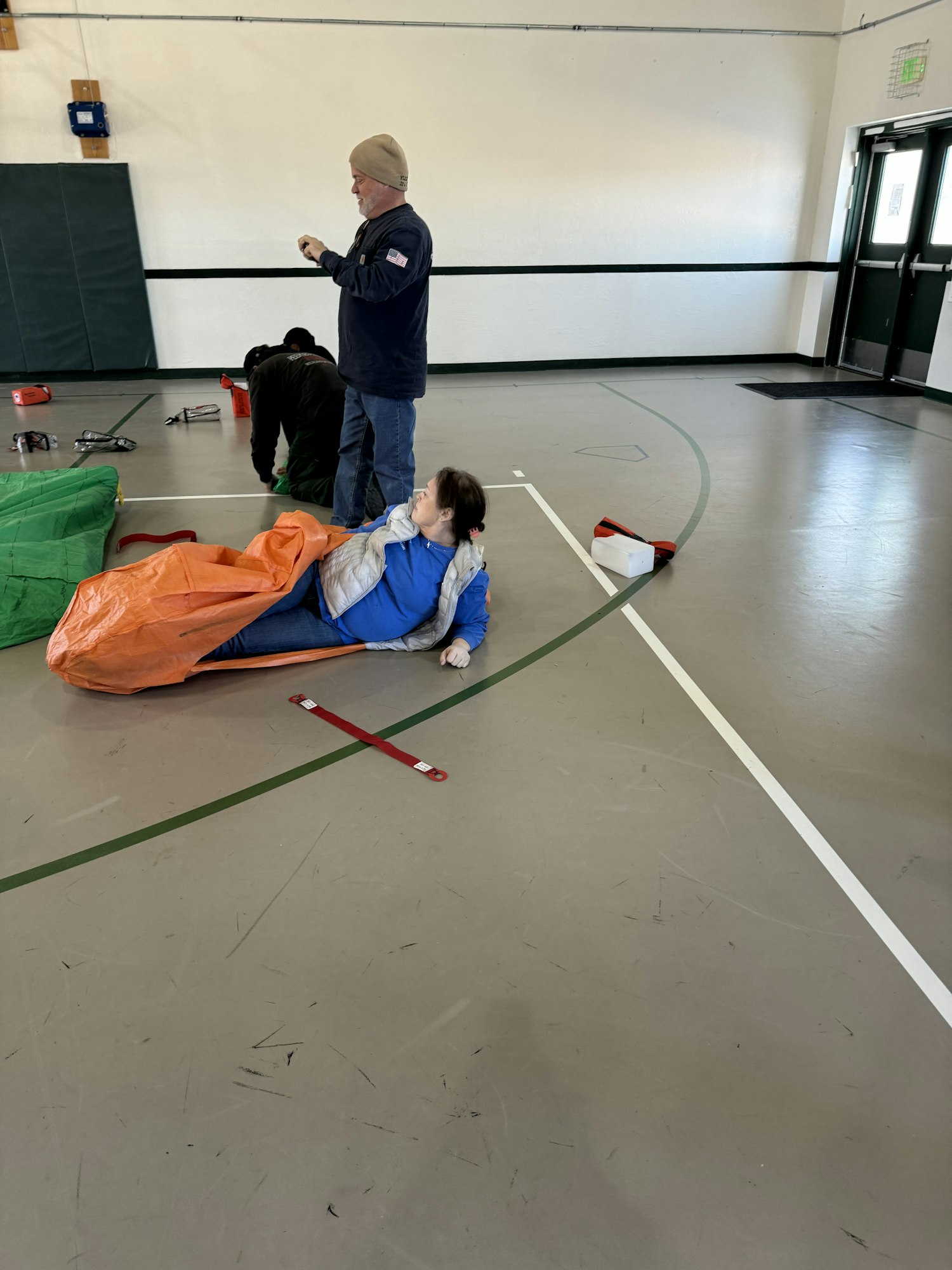 A person maneuvers a sled in a gym with another person assisting. Various items and equipment are scattered on the floor.