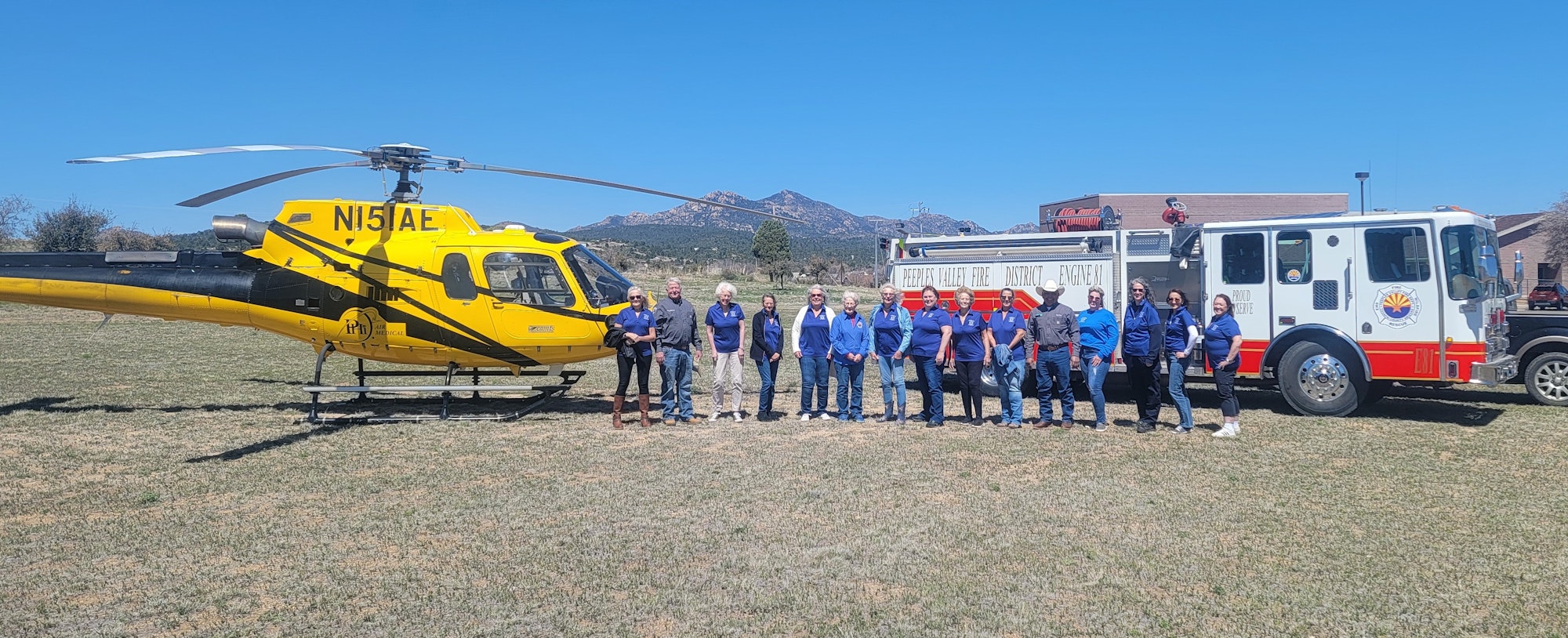 A group of people stand between a yellow medical helicopter and a fire engine under clear skies.