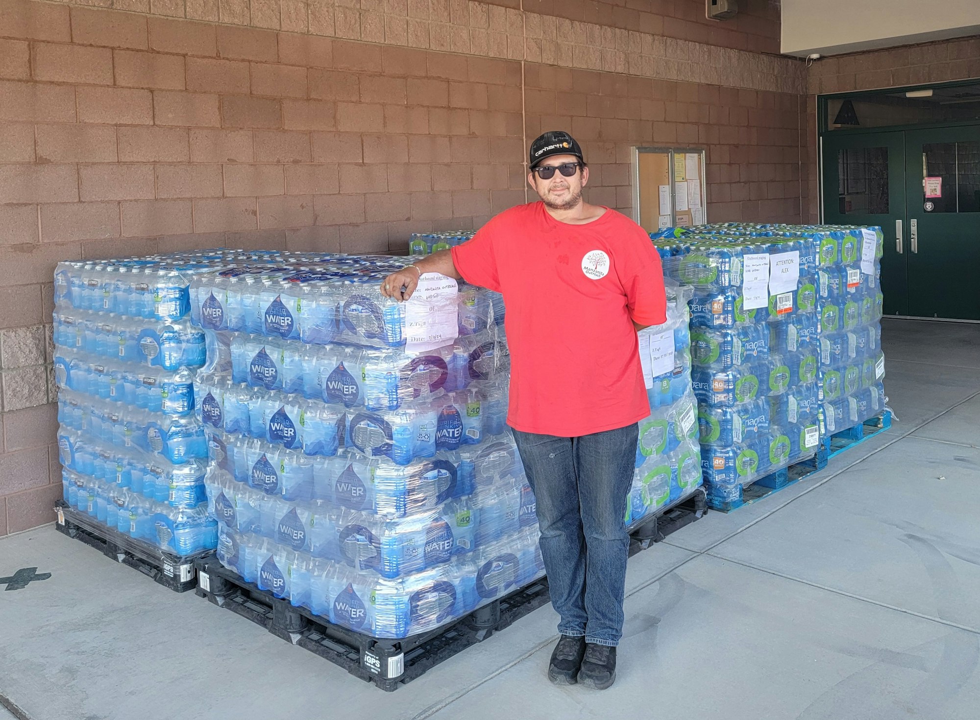 Man stands next to stacked water bottle packs outside a building.