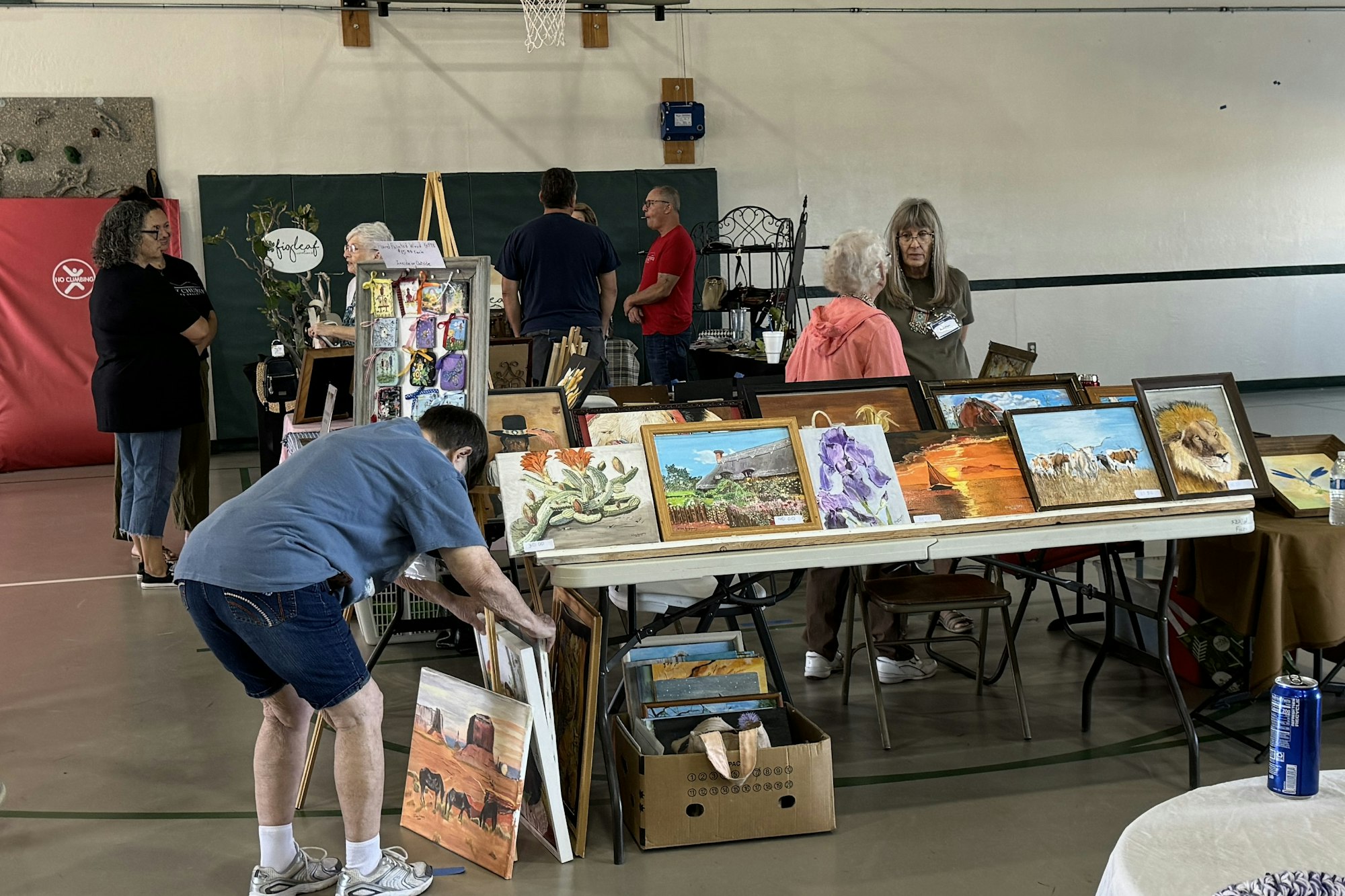 An indoor art fair with people browsing tables displaying paintings.