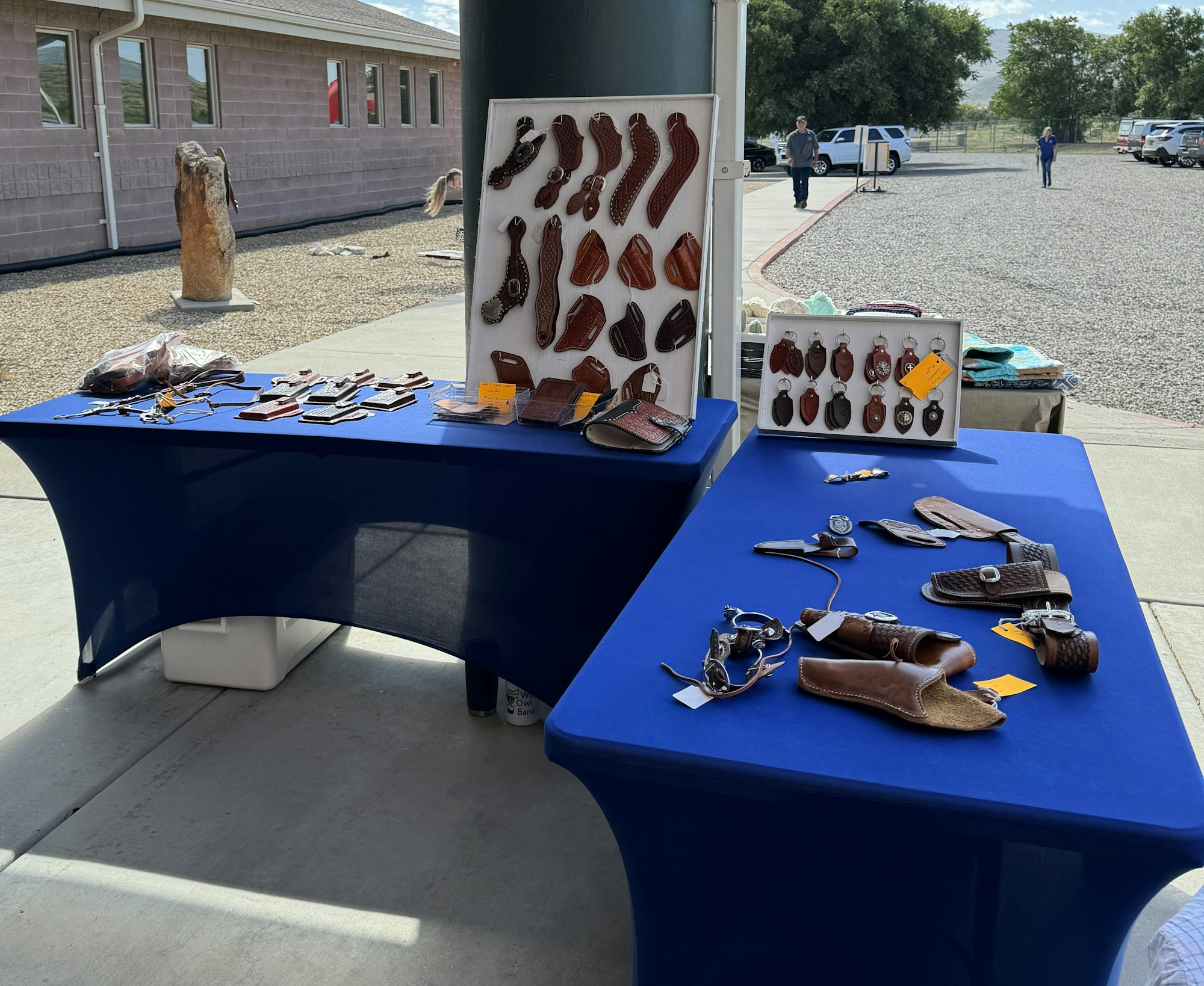 Outdoor market stall with leather goods, watches on display, people in the background, sunny day.