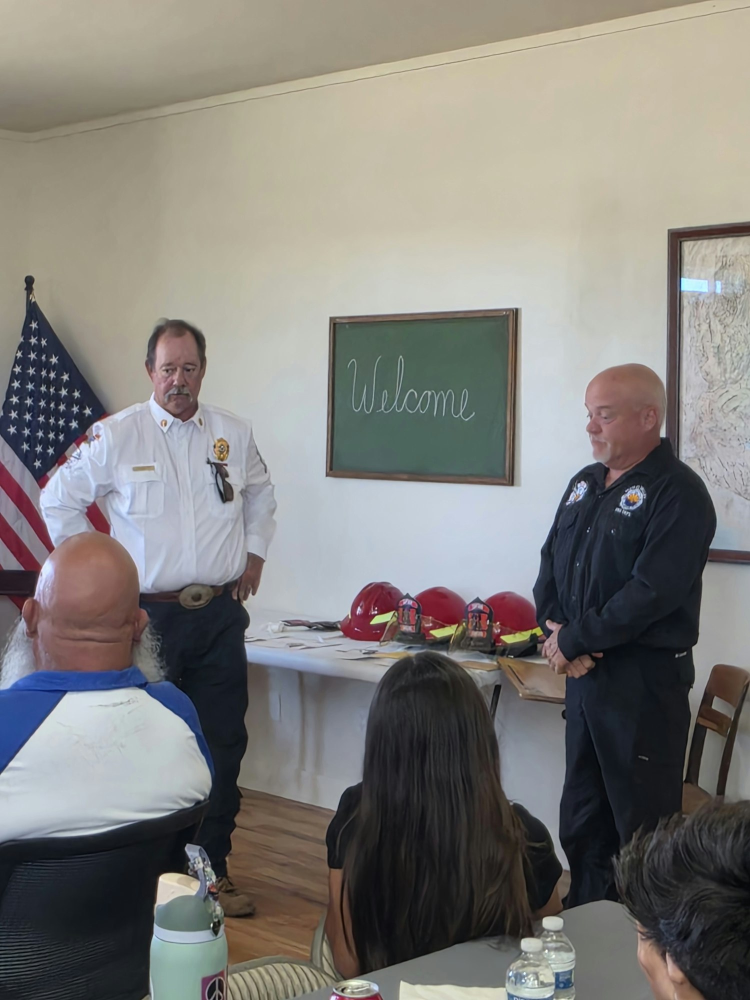 A welcome event featuring two men in uniform speaking, with a chalkboard, an American flag, and fire hats on a table.