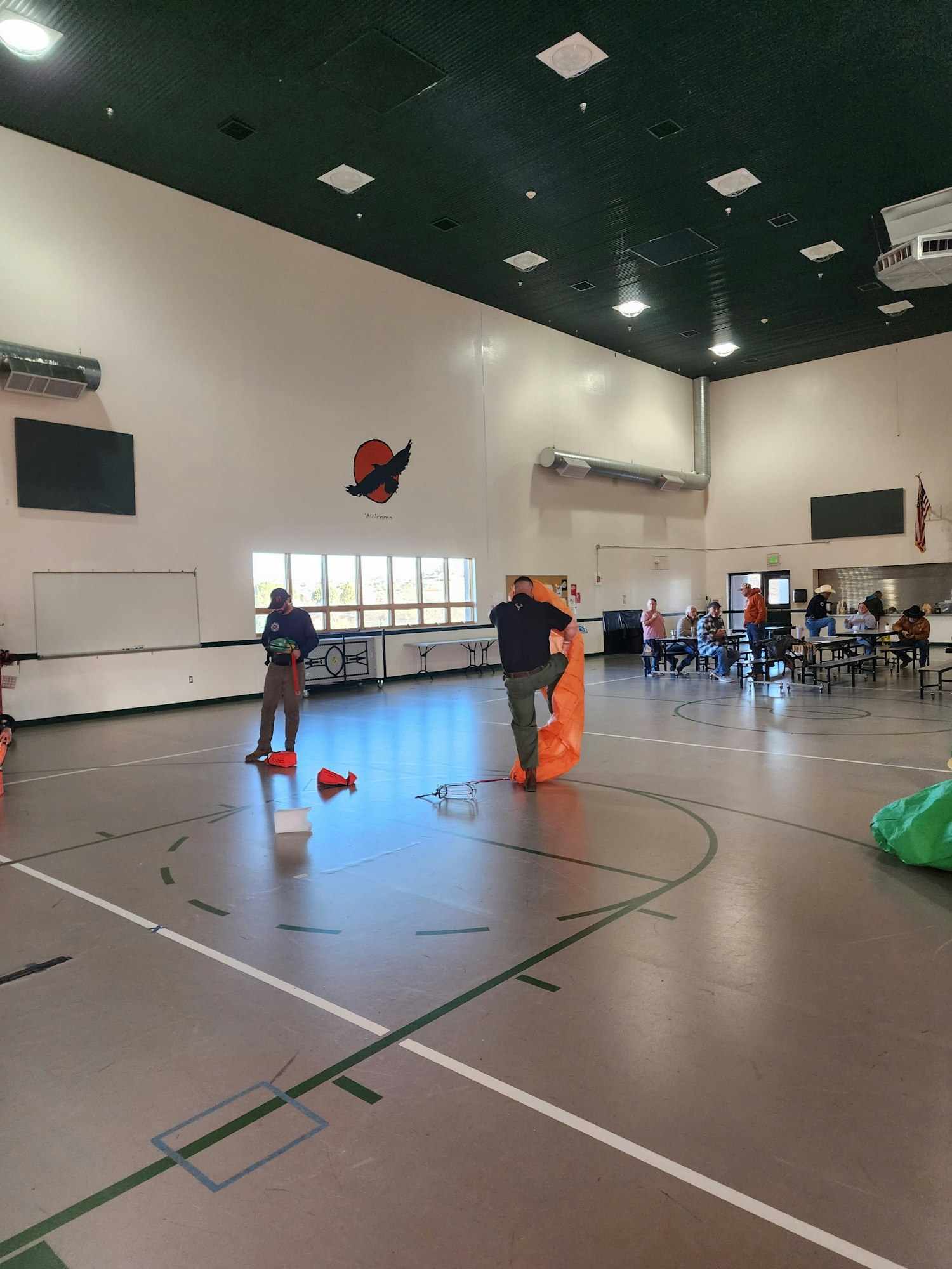 An indoor gym with green flooring, people preparing equipment, and tables in the background.