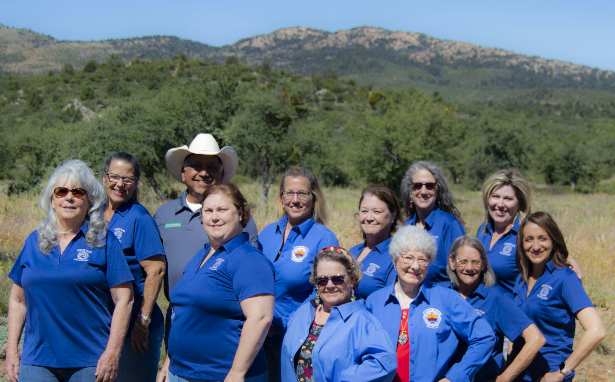A group of people in matching blue shirts posing outdoors with trees and mountains in the background.