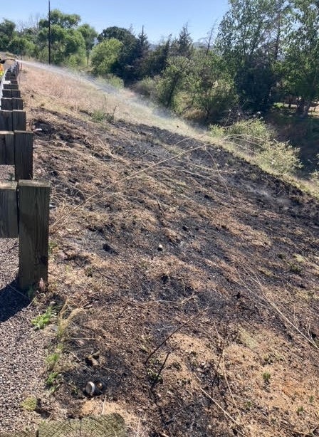 Scorched ground beside a wooden fence with some smoke, indicating a recent small fire or controlled burn.