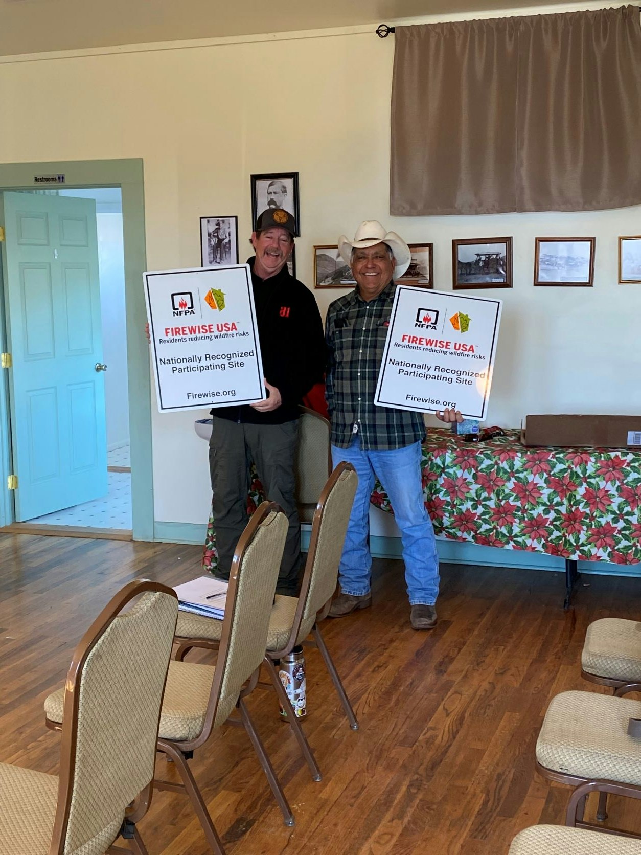 Two people holding signs with logos and text inside a room with chairs and pictures on the wall.