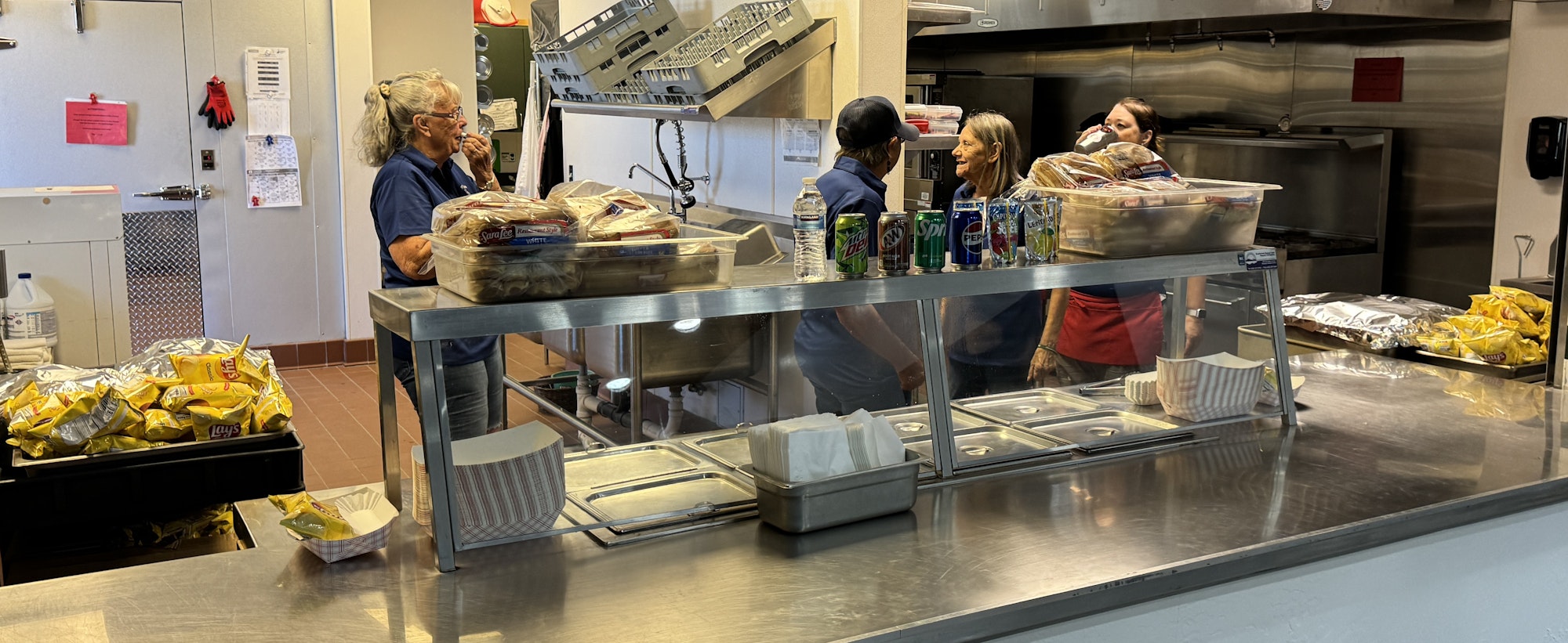 A cafeteria serving area with workers, packed chips, drinks, and bread visible behind glass counters.