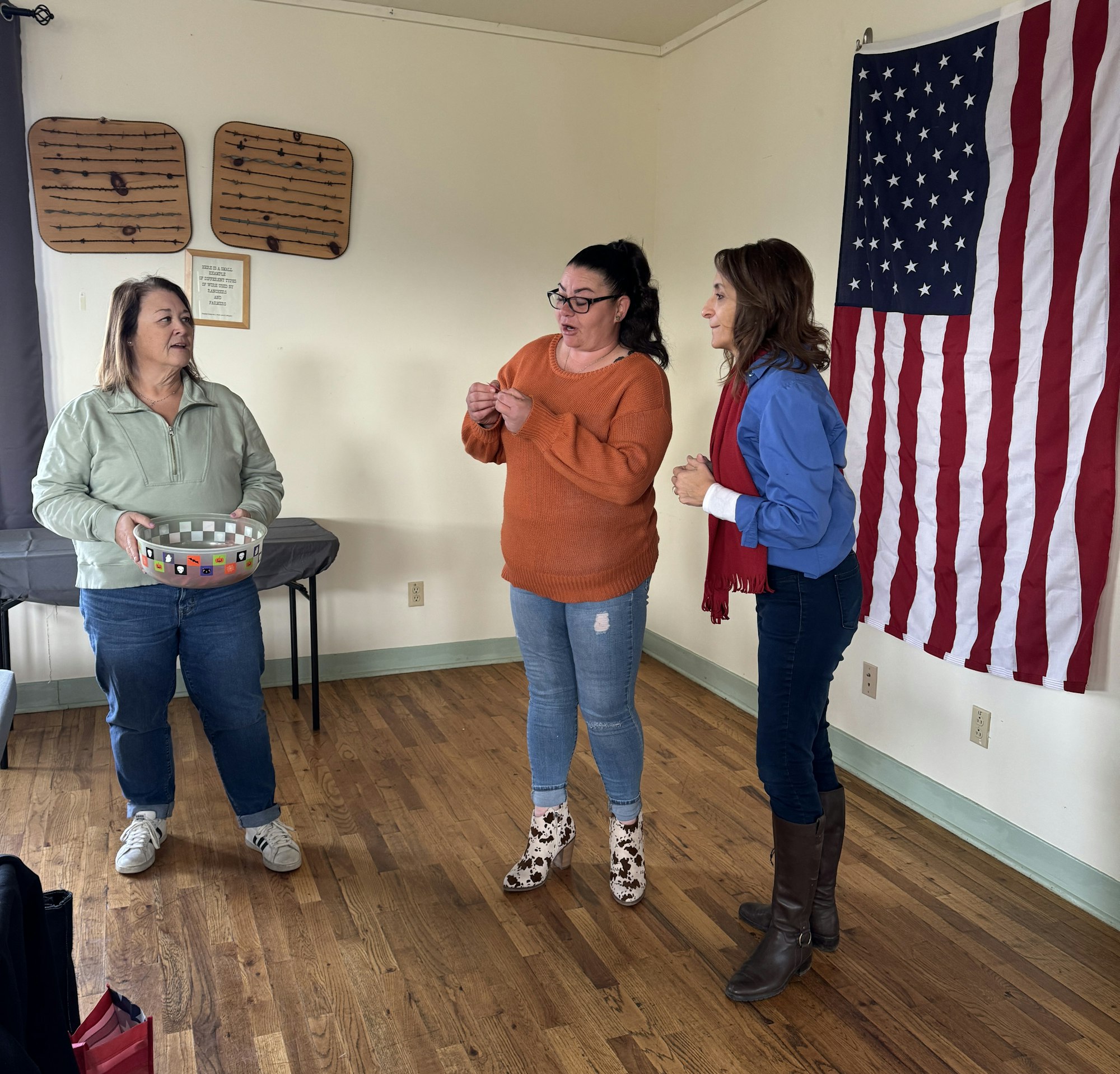 Three women chatting in a room with an American flag and two wooden message boards on the wall.