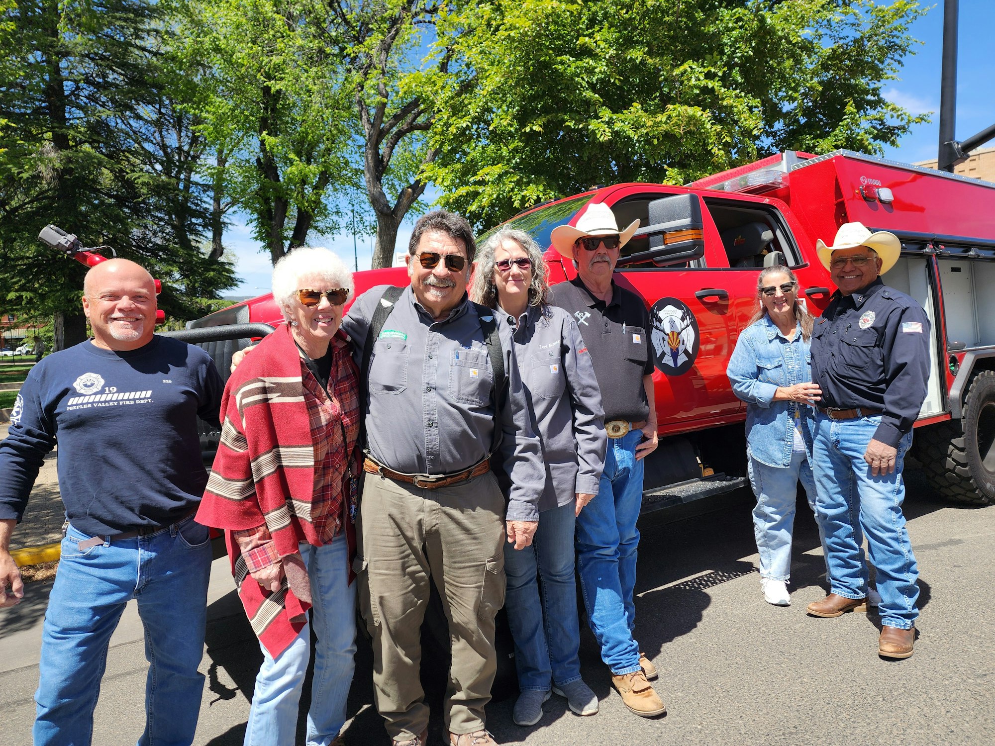 A group of six people, outdoors, posed near a red vehicle, smiling and dressed casually, surrounded by trees.