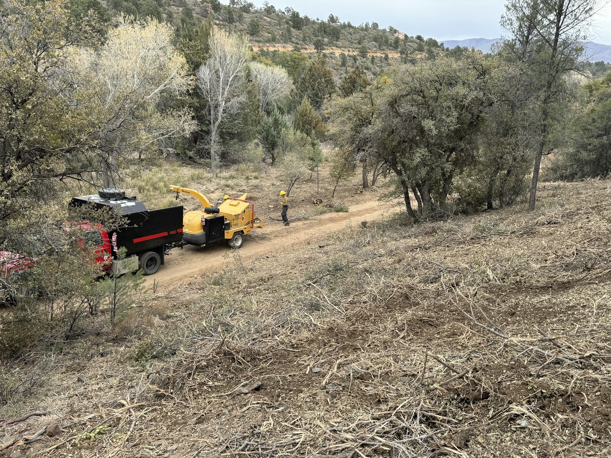 A red truck and a yellow wood chipper are on a dirt path in a wooded area, with a person in a hard hat working nearby.