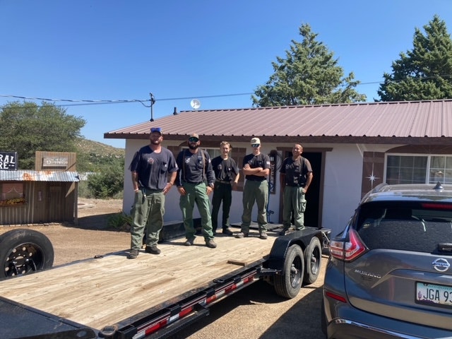 Five men standing on a flatbed trailer in front of a building with a car parked nearby.