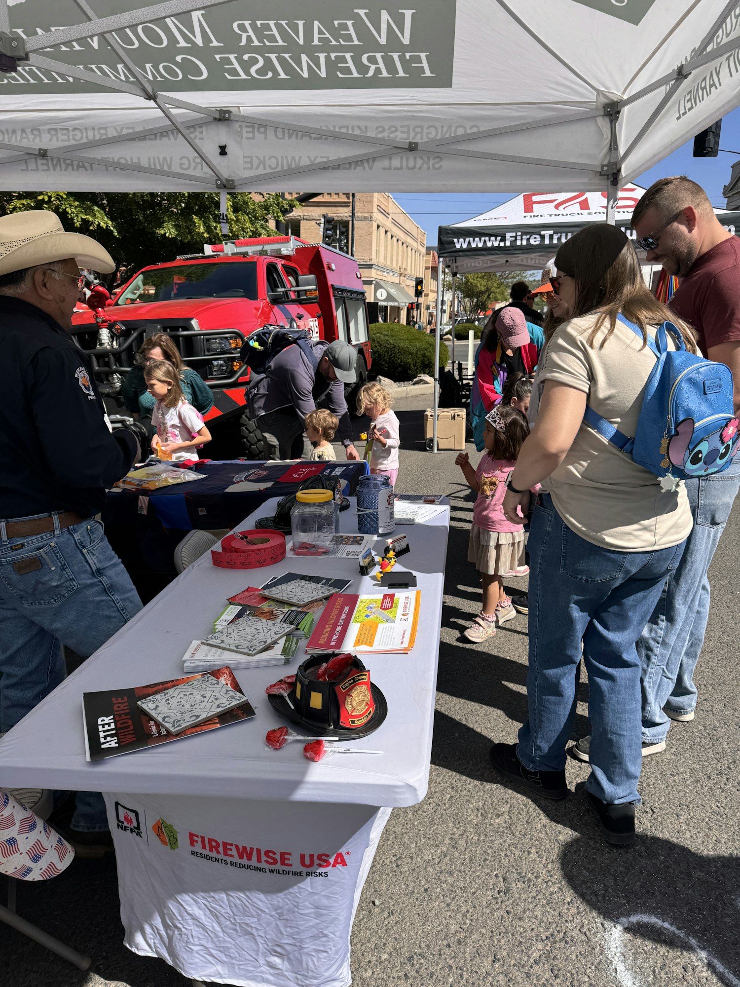 People gather around a booth at an outdoor event, with items like fire safety materials and a red fire truck in the background.