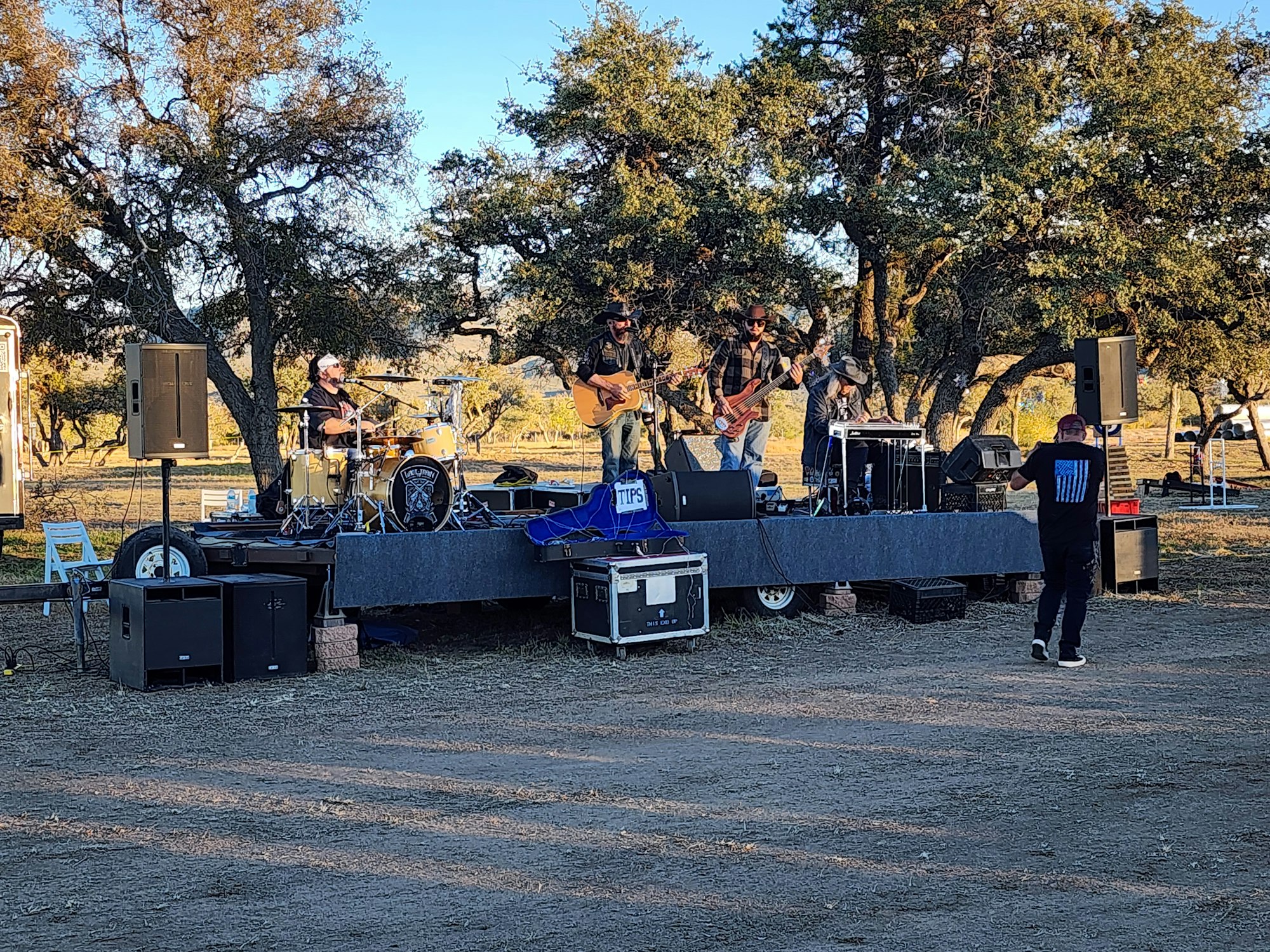 A band performs on an outdoor stage surrounded by trees, with speakers and a drum set visible. A man stands in front of the stage.
