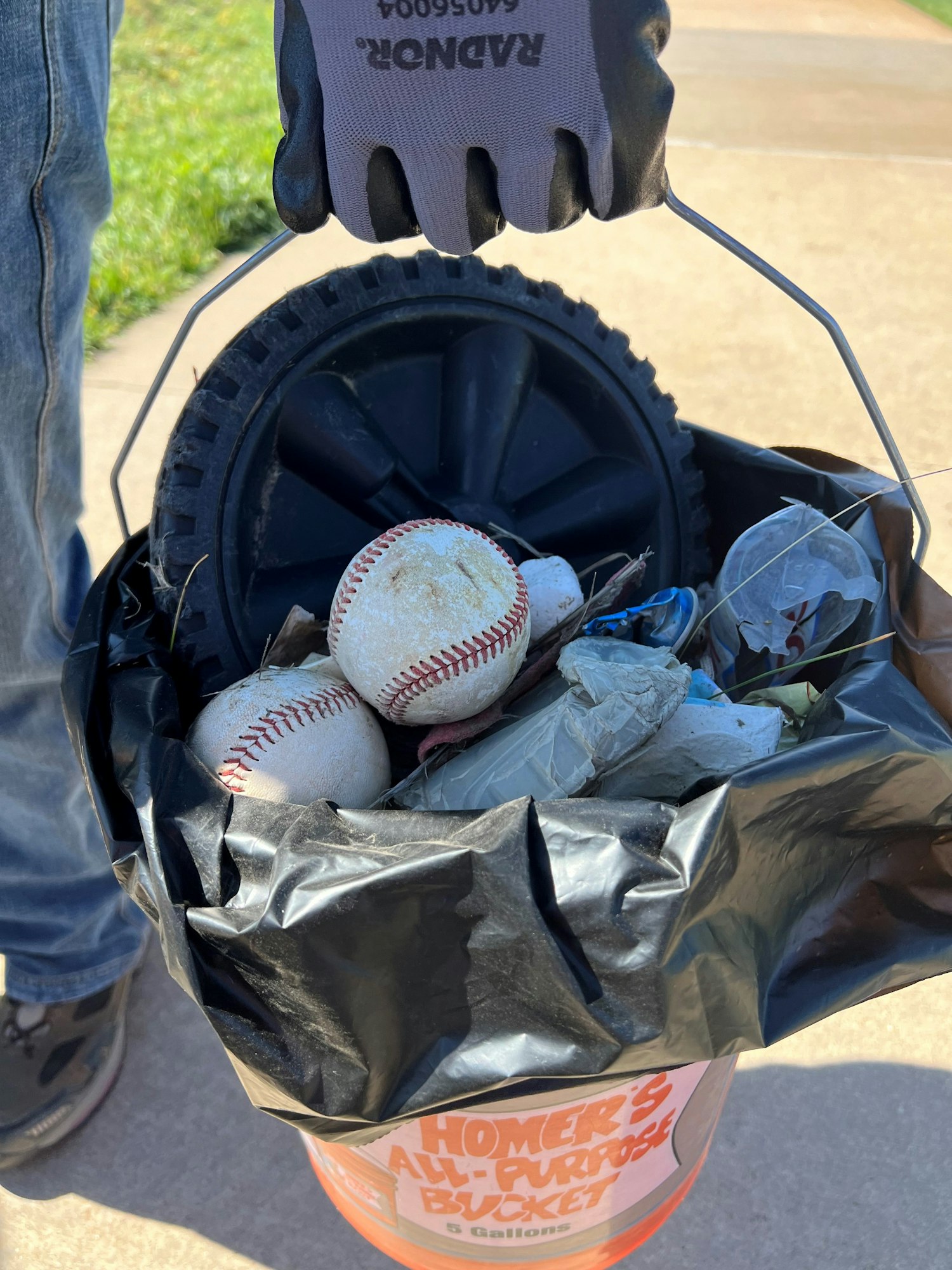 A gloved hand holding a bucket filled with trash, including baseballs and a wheel, lined with a black trash bag.