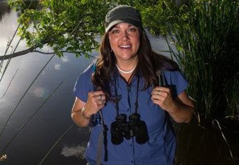 A smiling woman stands by a body of water, wearing a cap and holding binoculars, ready for outdoor exploration.