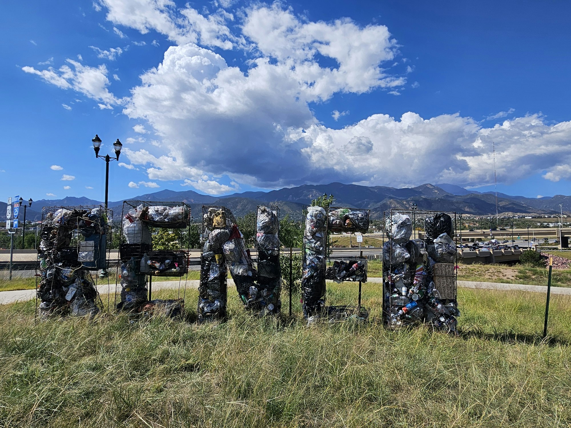 Large letters spelling "RENT" made from trash encased in wire, set against a mountainous landscape and blue sky with clouds.