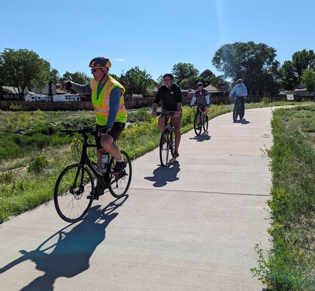 Four cyclists on a path, one wearing a high-visibility vest, with a pedestrian in the background under a sunny sky.