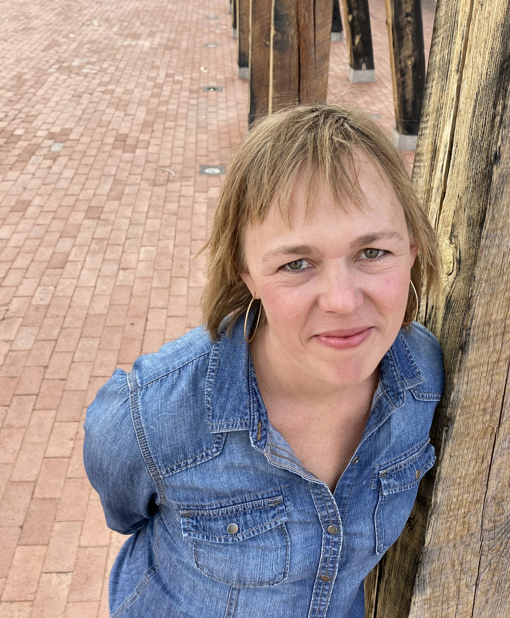The image shows a woman wearing a denim shirt, leaning against a wooden post on a brick pathway.