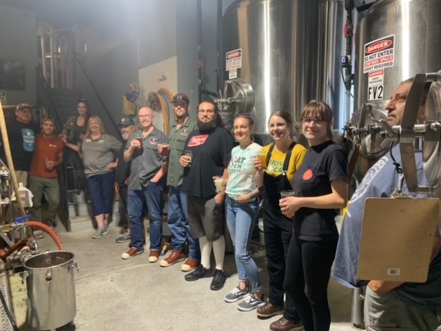 A group of people gathered in a brewery, holding drinks and posing for a photo, with brewing equipment in the background.