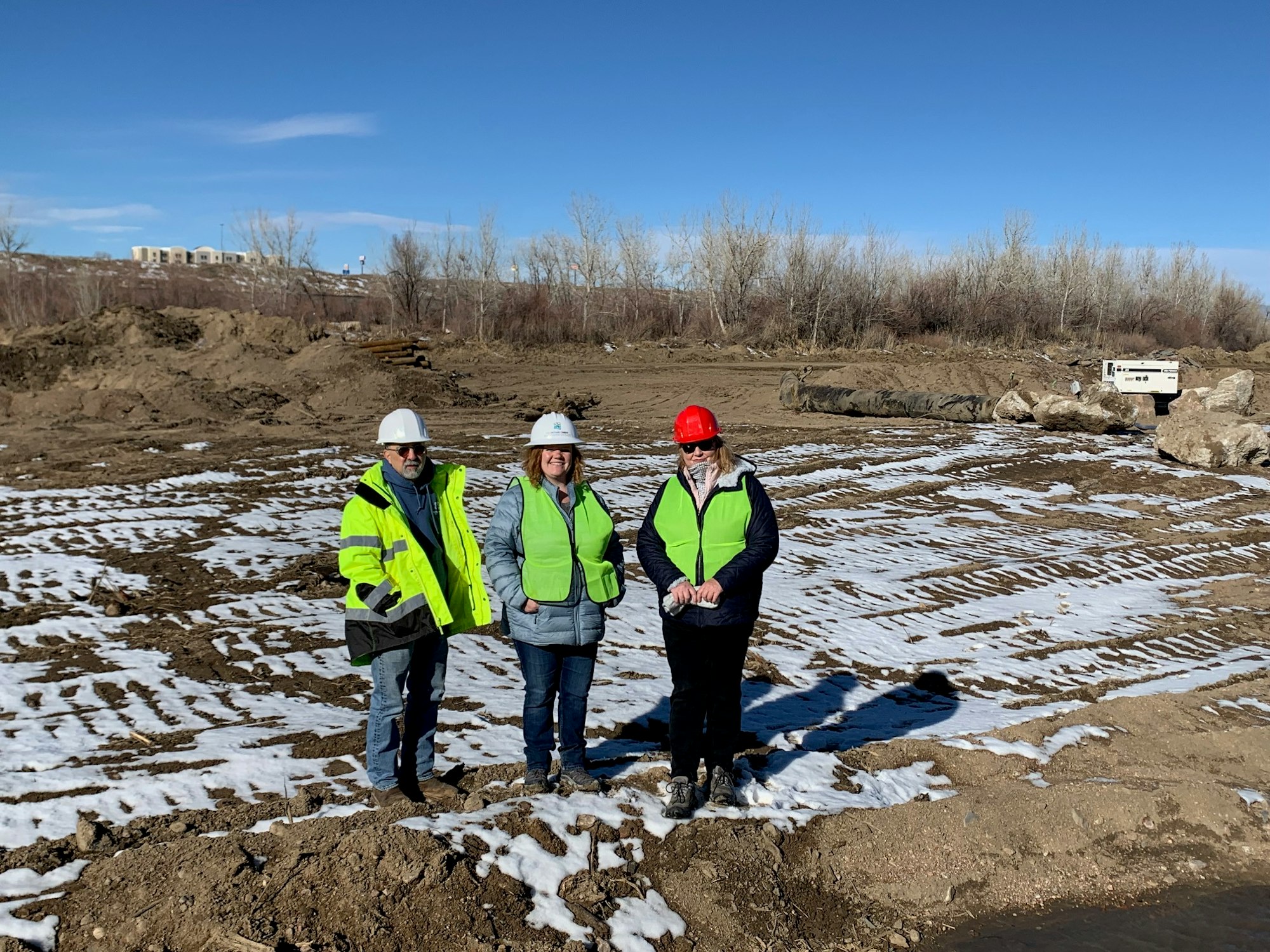 Three people in hard hats and high-vis vests stand on a snowy construction site.