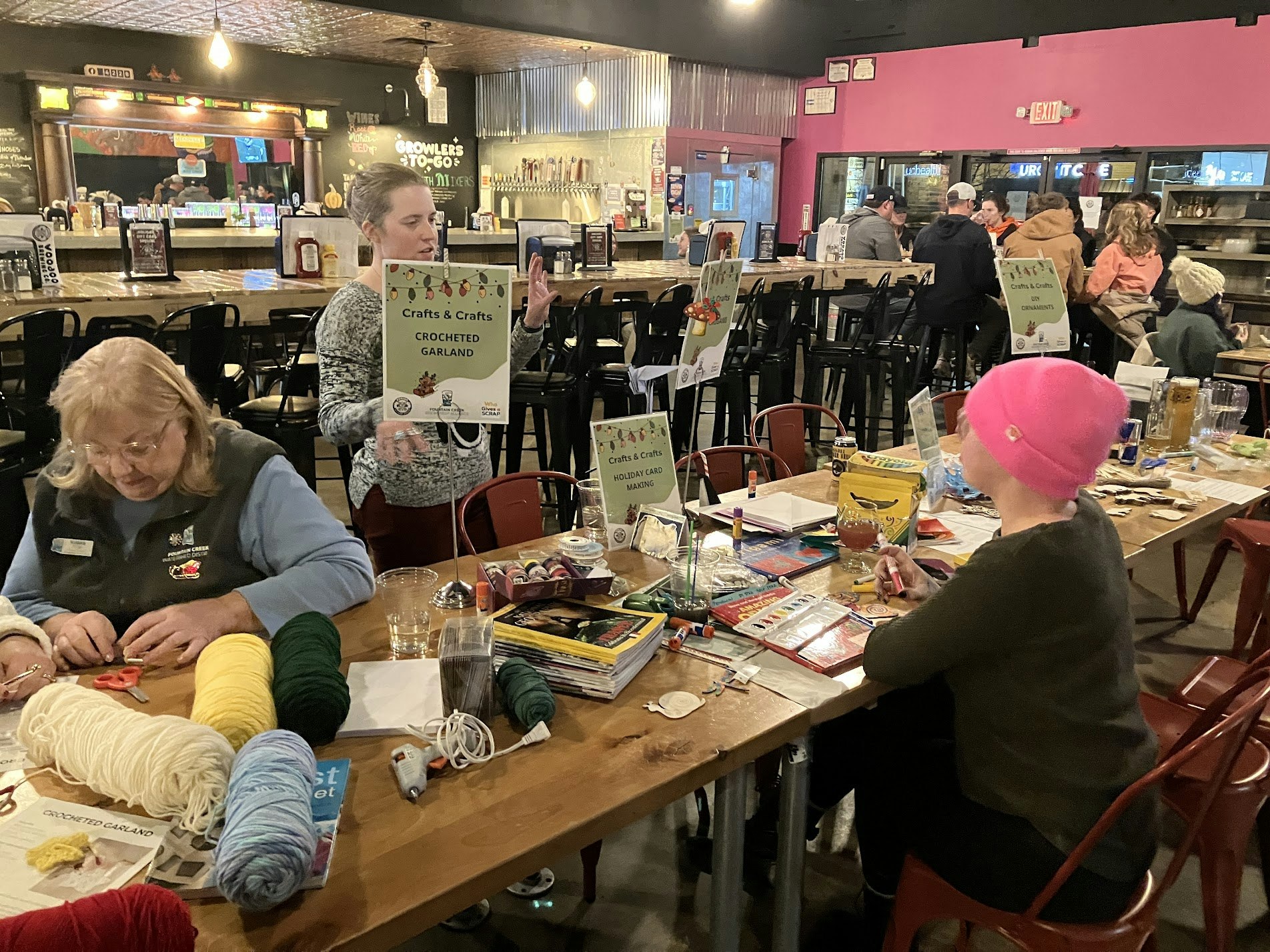 A craft event at a bar with people making items like crocheted garlands and greeting cards, surrounded by supplies and drinks.